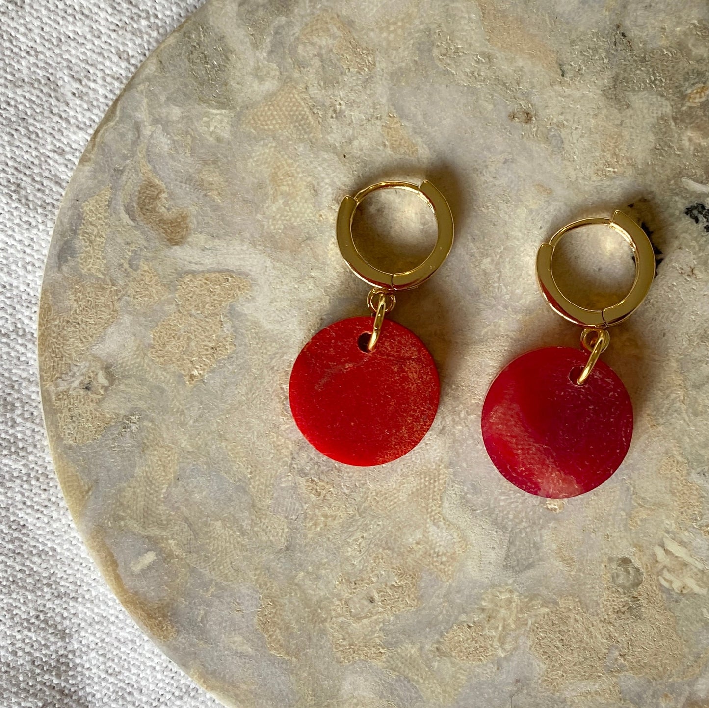 Gold hoop earrings with red circular charms on a stone surface